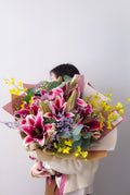 A person in a cream sweater holds a large, beautifully wrapped flower bouquet featuring pink and white lilies, yellow wildflowers, and green foliage against a plain light gray background.